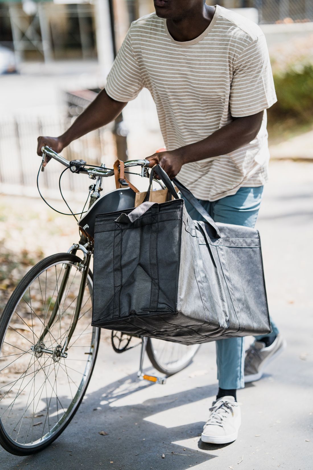 A rider pushing a bicycle with a large insulated delivery bag hanging from the handlebars