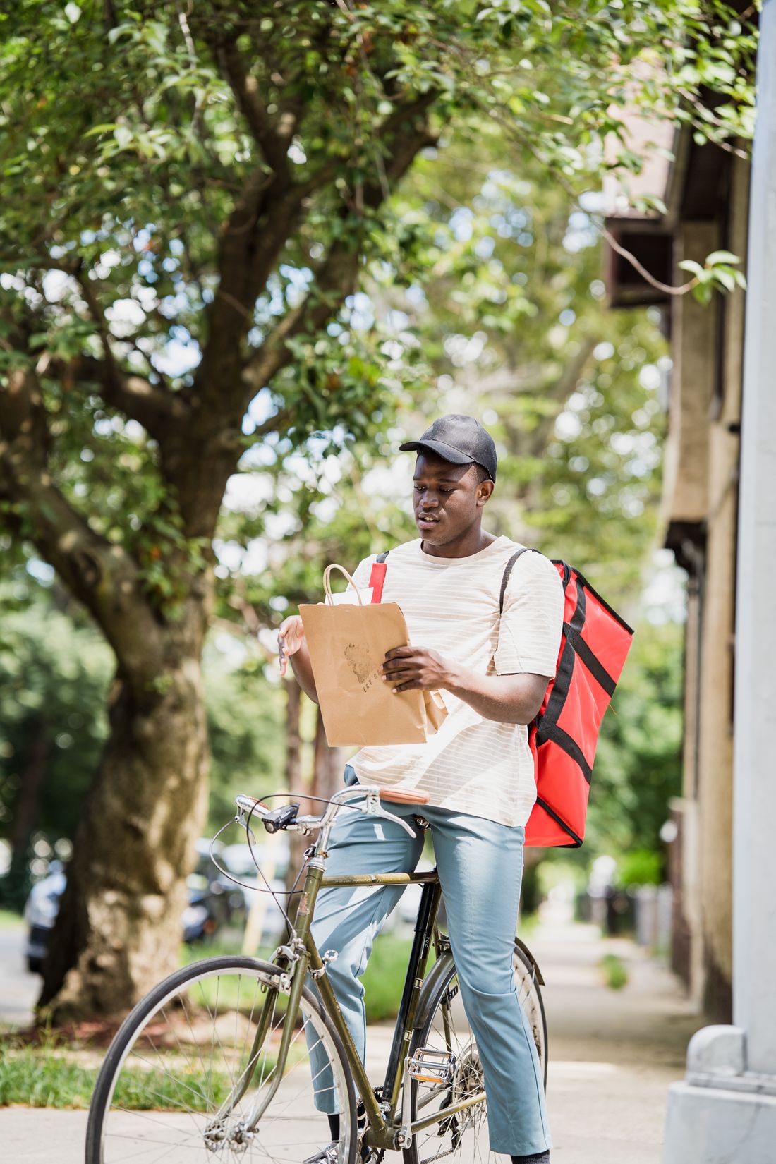 A delivery rider with a red backpack checking a paper-bag order beside his bicycle