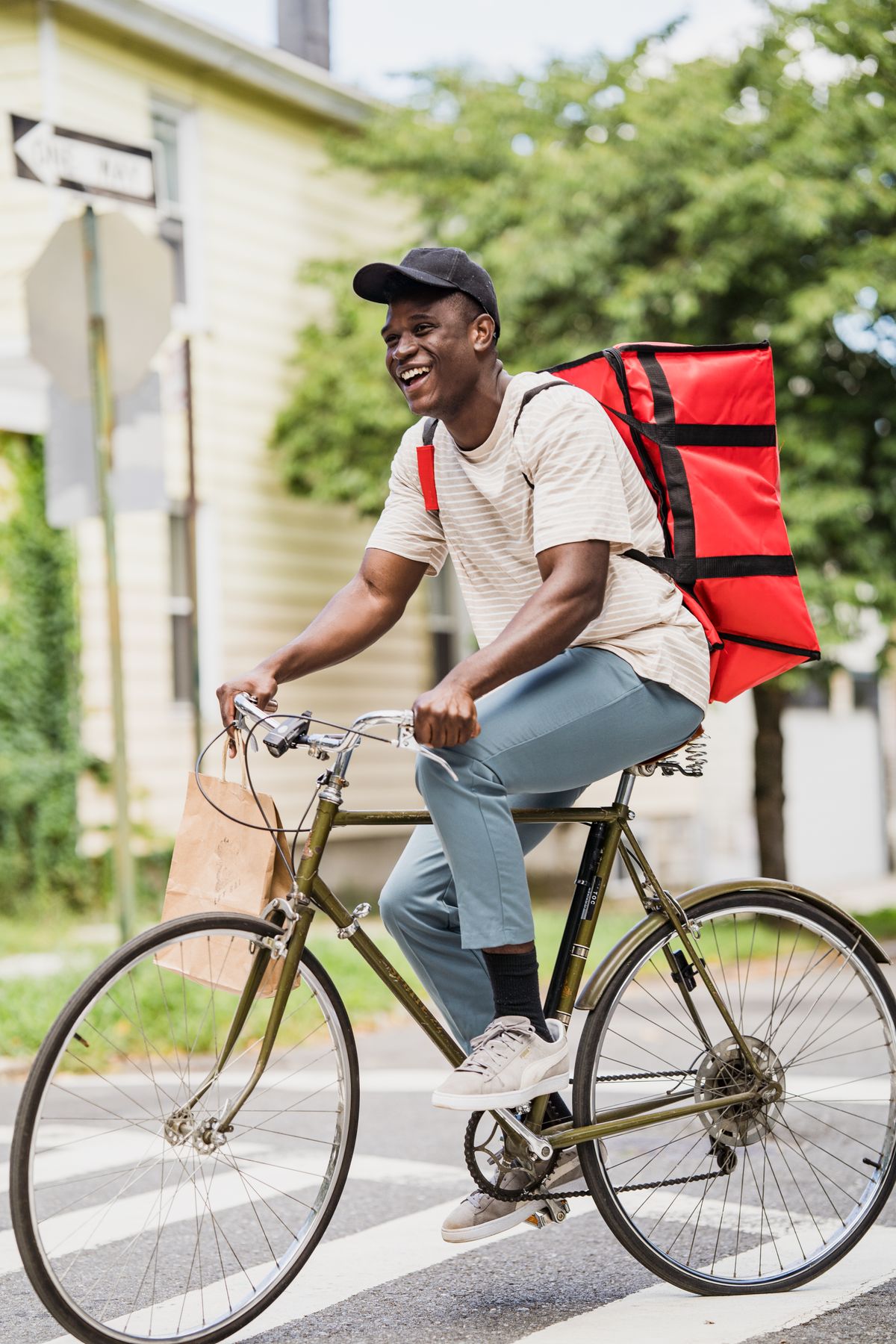 A smiling delivery rider in a cap, cycling with a red delivery backpack and a paper bag on the handlebars
